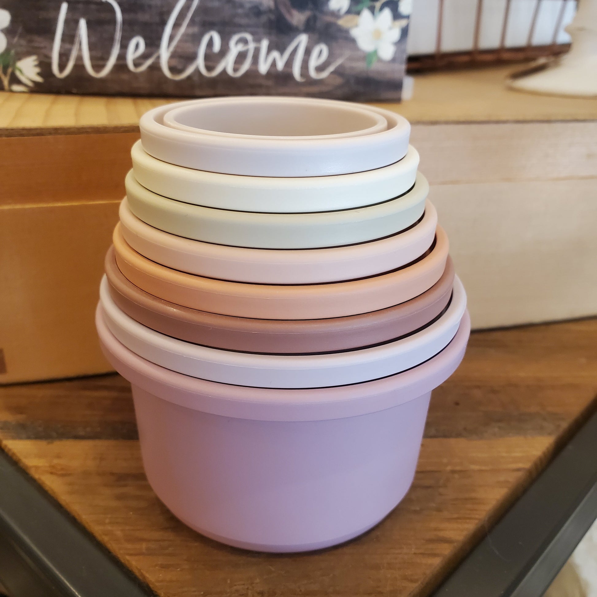 A stack of various sized beige and pink ceramic cups and mugs on a wooden surface with a 'Welcome' sign in the background.