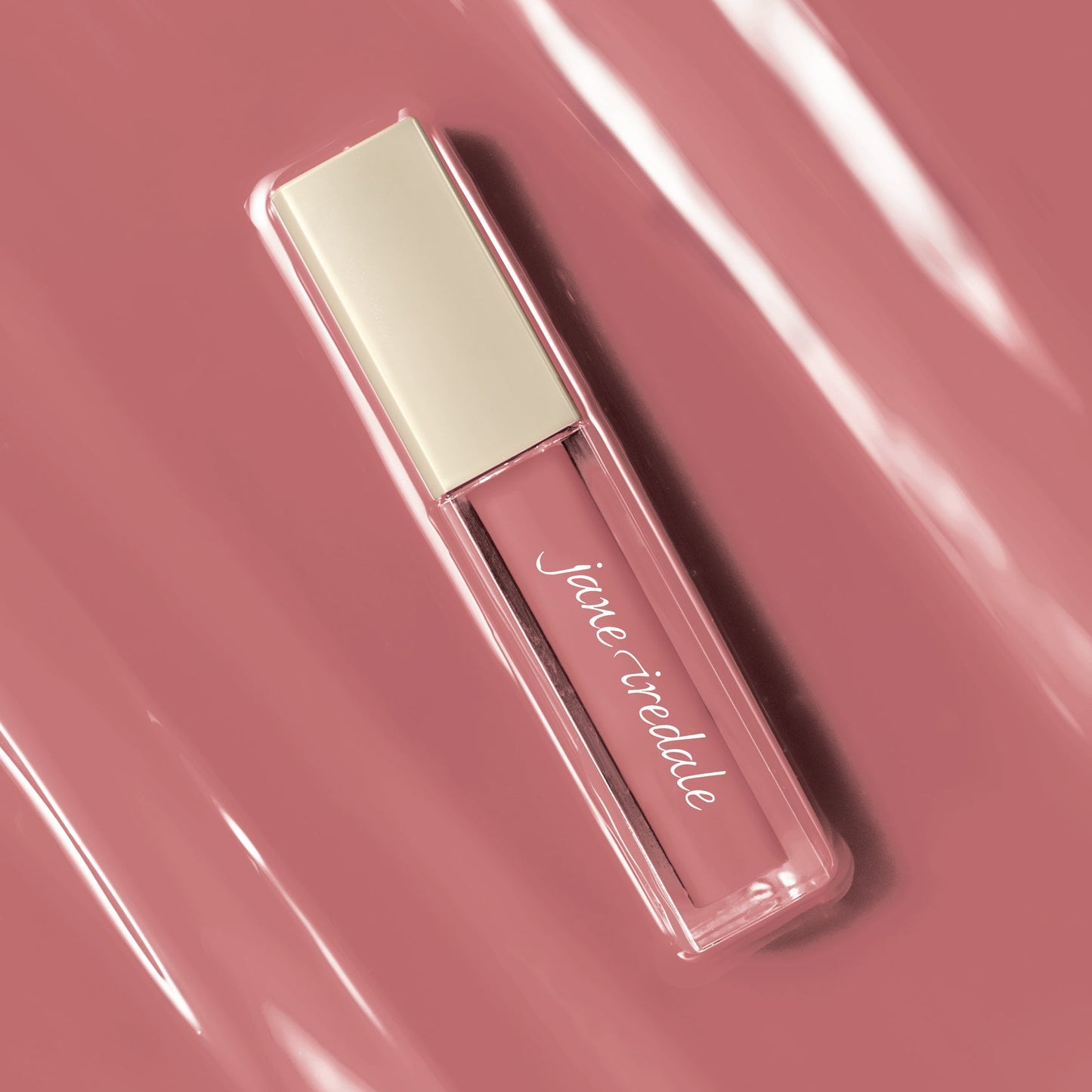 A rectangular liquid lipstick tube by Jane Iredale, filled with a dusty rose pink gloss and featuring a pale cream cap, set against a matching dusty rose background with glossy reflections.