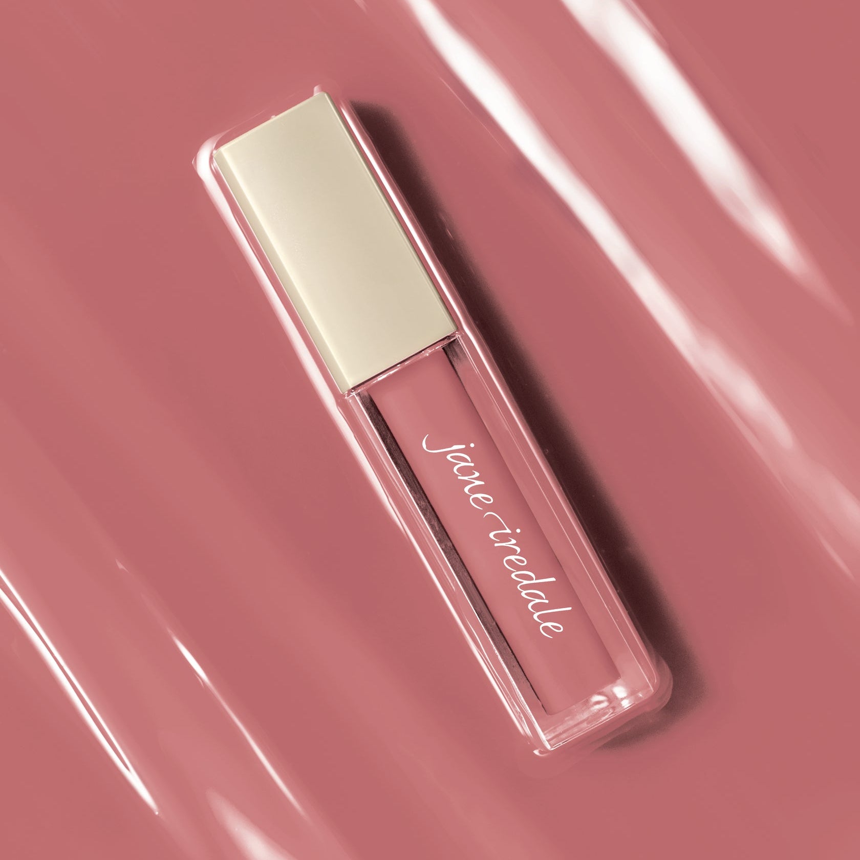 A rectangular liquid lipstick tube by Jane Iredale, filled with a dusty rose pink gloss and featuring a pale cream cap, set against a matching dusty rose background with glossy reflections.